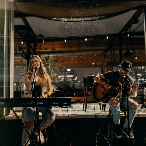 A woman sings at a microphone while a man plays guitar beside her on a stage with city lights in the background.