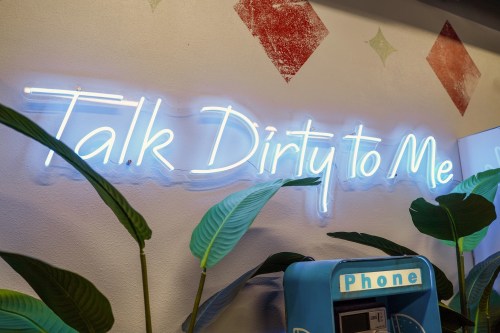 Neon sign reading "Talk Dirty to Me" with green plants and a vintage phone booth in the background.