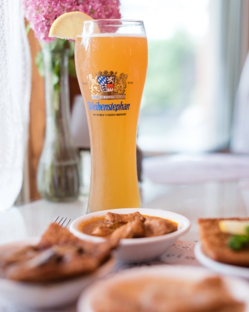 A tall glass of Weihenstephan beer with a lemon slice, surrounded by dishes of food and a vase of pink flowers.