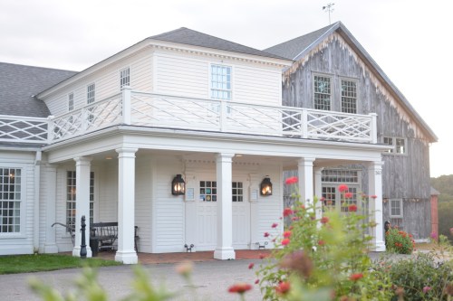 A charming white house with a porch and balcony, surrounded by colorful flowers and a rustic wooden building nearby.
