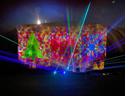 Colorful light projections of trees and patterns illuminate a mountain, with vibrant lasers cutting through the night sky.