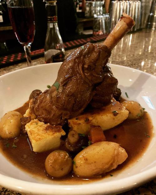 A plated dish featuring a braised lamb shank on a bed of vegetables and potatoes, garnished with herbs.