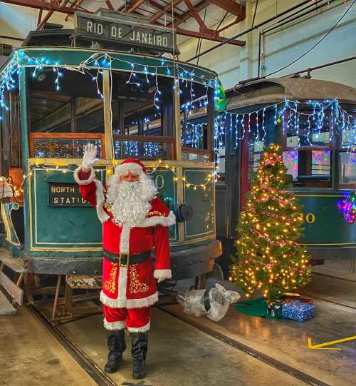 A man in a Santa costume waves beside a decorated vintage tram and a Christmas tree with lights.