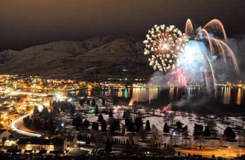 Colorful fireworks illuminate a snowy landscape and a lakeside town at night, with mountains in the background.
