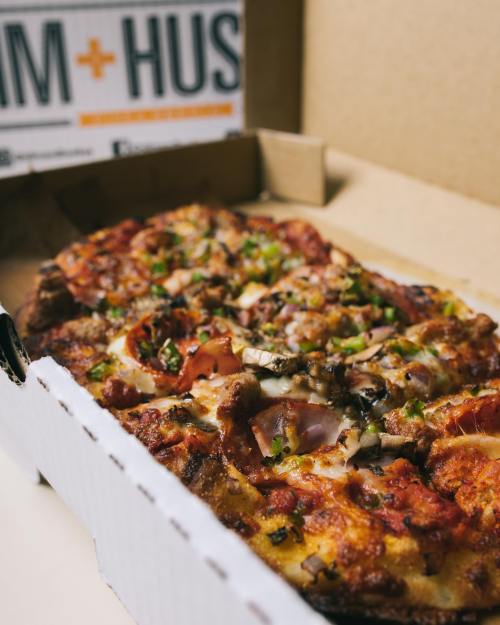 A close-up of a pizza in a takeout box, topped with various meats, vegetables, and melted cheese.