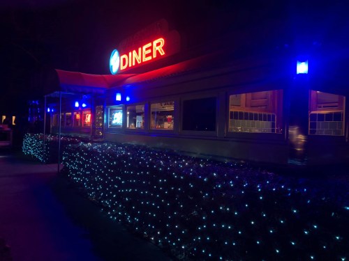 A brightly lit diner at night, featuring blue lights and a glowing sign, surrounded by decorative lights.