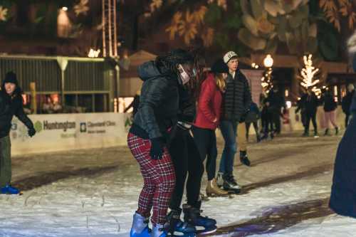 A group of people ice skating at a festive outdoor rink, surrounded by holiday decorations and lights.