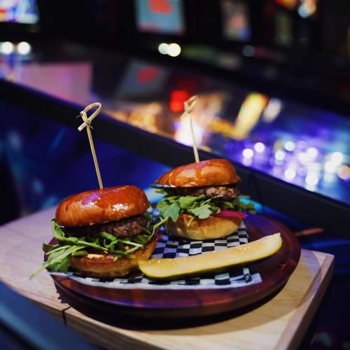 Two gourmet burgers on a wooden platter, garnished with greens and pickles, set against a colorful, blurred background.