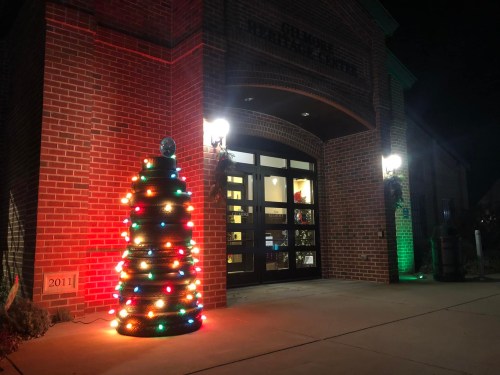 A brightly lit Christmas tree made of lights stands outside a brick building at night, with festive decorations visible.
