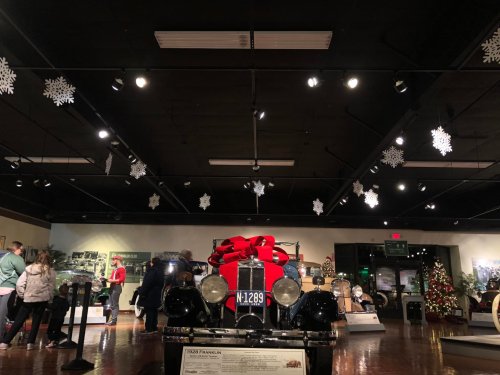 A vintage car with a large red bow is displayed in a festive museum setting, decorated with snowflakes and holiday decor.
