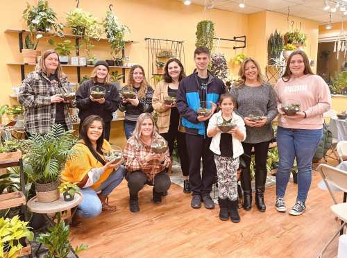 A group of ten people, including children, holding potted plants in a cozy plant shop setting.