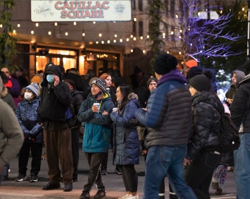 A crowd of people in winter clothing gathers in Cadillac Square, enjoying the festive atmosphere with lights and decorations.