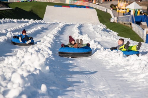 Three children slide down a snowy hill on inflatable tubes, enjoying a winter day at a snow park.