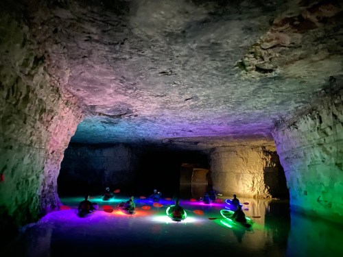 A group of kayakers with glowing lights paddles through a colorful, illuminated cave.