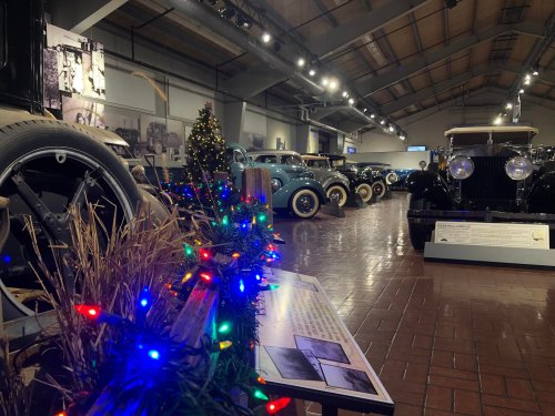 A vintage car museum decorated with holiday lights and a festive display, showcasing classic cars in the background.