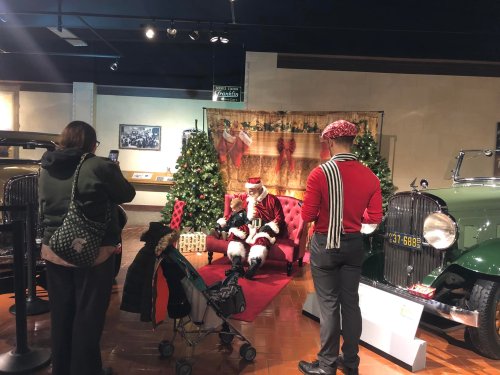 A Santa Claus sits on a red couch in a museum, surrounded by Christmas trees, while visitors take photos.