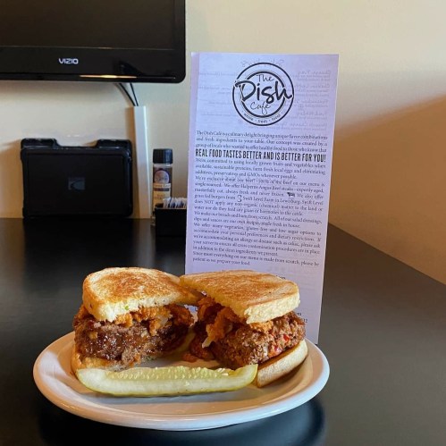 A plate with a sandwich made of fried chicken between two slices of bread, accompanied by a pickle and a menu in the background.