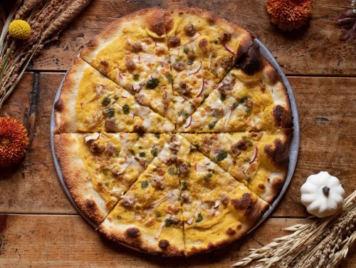 A round pizza topped with cheese, green peppers, and slices of apple, cut into eight pieces, on a wooden table.