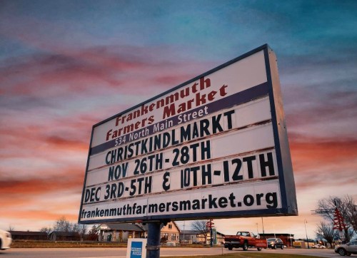 Sign for Frankenmuth Farmers Market with event dates for Christkindlmarkt and website information against a colorful sky.