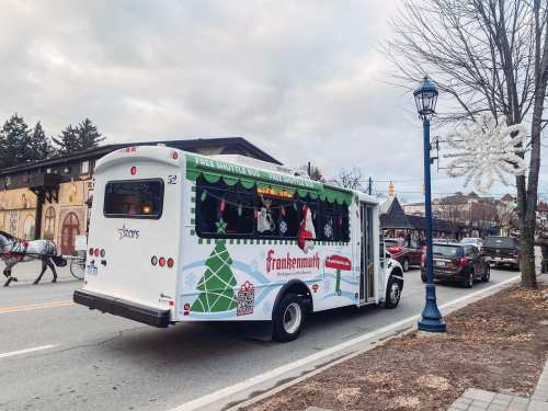 A festive shuttle bus decorated for the holidays, parked on a street with winter decorations and a horse-drawn carriage nearby.