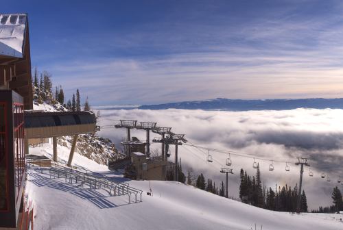 A snowy mountain landscape with ski lifts and a lodge, surrounded by clouds and distant mountains under a clear sky.