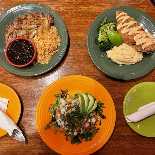 A table with three colorful plates: grilled steak with onions, fried chicken, and a fresh salad with avocado.