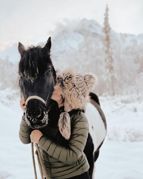 A person in a furry hat kisses a black and white horse in a snowy landscape with mountains in the background.