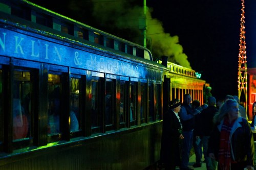 A vintage train station at night, with steam rising and people gathered near a brightly lit train.