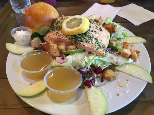 A plate with grilled salmon topped with lemon, served on a bed of salad, croutons, apple slices, and two cups of dressing.