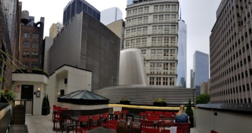 A rooftop terrace with red chairs and tables, surrounded by tall buildings and a cloudy sky.