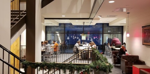 A cozy café interior with people seated at tables, large windows, and festive decorations along the railing.