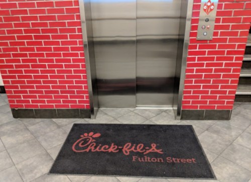A Chick-fil-A welcome mat in front of an elevator, with a red brick wall in the background.
