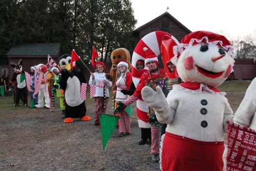 A lineup of festive characters in costumes, including a snowman, gingerbread man, and candy cane, holding flags and decorations.