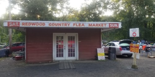 A small red building with a sign reading "Redwood Country Flea Market" surrounded by parked cars and trees.