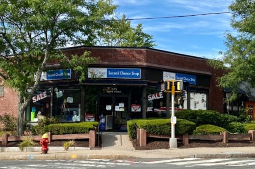 A storefront of the "Second Chance Shop" with a sale sign, surrounded by greenery and a clear blue sky.