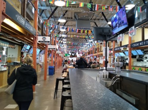 A bustling food hall with flags hanging, featuring various food stalls and patrons dining at tables.