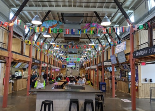 A vibrant food hall with colorful flags, wooden stalls, and people enjoying the space. Bright and welcoming atmosphere.