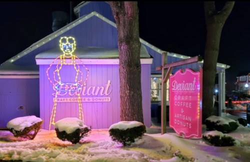 A brightly lit sign of a figure stands outside a coffee and donut shop, surrounded by snow and festive decorations.