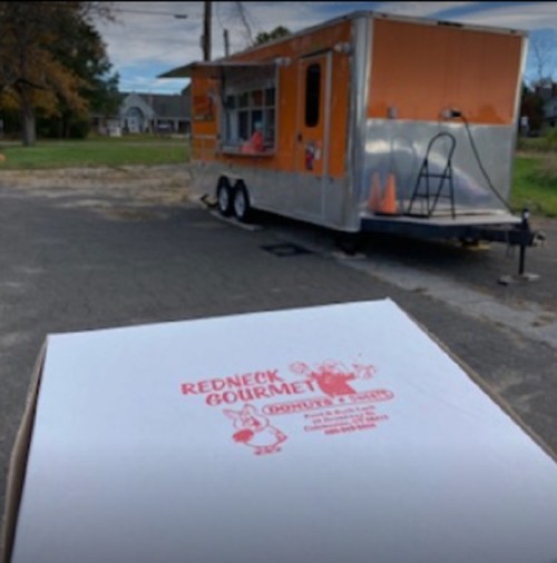 A food truck with an orange exterior parked in a lot, next to a large takeout box labeled "Redneck Gourmet."