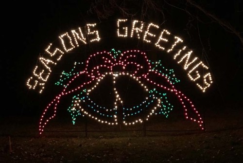 Colorful holiday lights display reading "Seasons Greetings" with bells and ribbons, set against a dark background.
