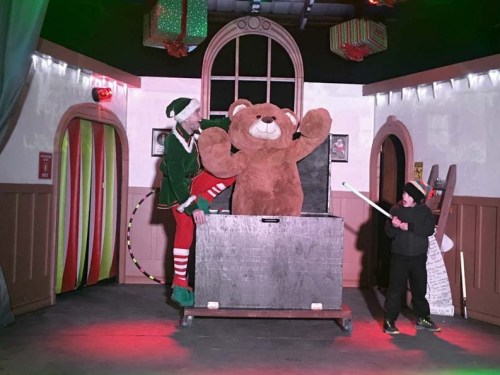 A performer in an elf costume interacts with a large teddy bear on stage, while another child holds a wand nearby.