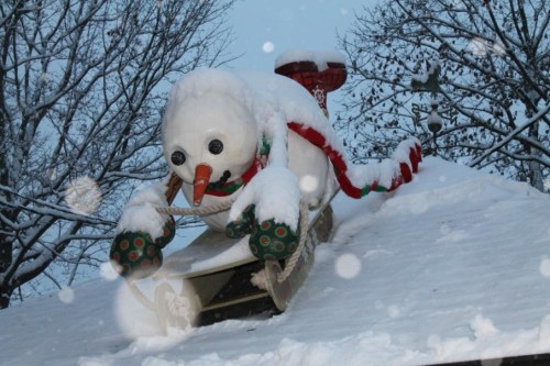 A snowman on a snowy roof, sliding down with a festive scarf and colorful gloves, surrounded by winter scenery.