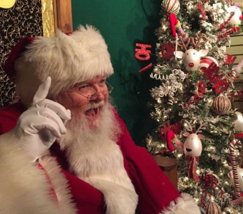 A cheerful Santa Claus in a red suit and hat, sitting beside a decorated Christmas tree, smiling and gesturing.