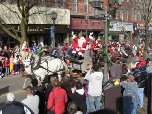 A festive parade scene with Santa on a horse-drawn carriage, surrounded by a cheering crowd of children and families.
