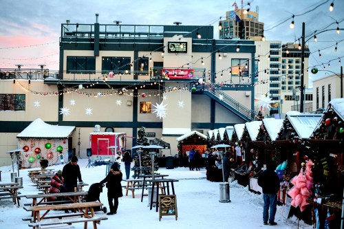 A festive outdoor market with holiday decorations, snow, and people enjoying the atmosphere under string lights.