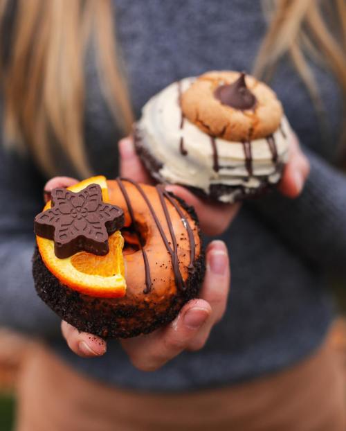 A person holds two decorated donuts: one with chocolate, orange, and a star-shaped cookie, and another with a chocolate chip.