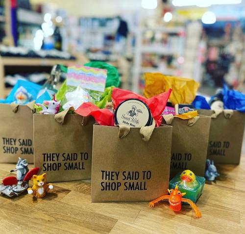 Brown paper bags filled with colorful gifts and toys, labeled "THEY SAID TO SHOP SMALL," arranged on a wooden surface.