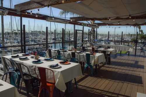A waterfront restaurant with tables set for dining, overlooking a marina filled with boats. Bright and inviting atmosphere.