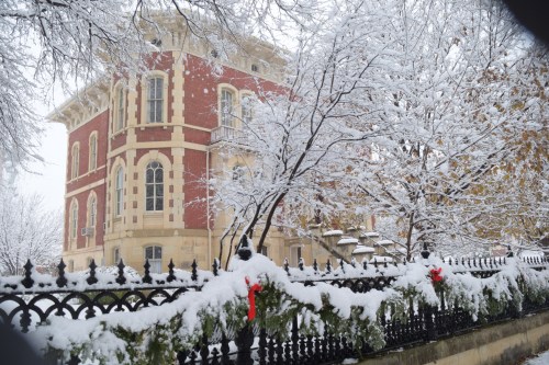 A historic red brick building surrounded by snow-covered trees and a decorated fence in a winter landscape.