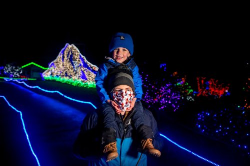A child smiles while riding on an adult's shoulders, surrounded by colorful holiday lights at night.